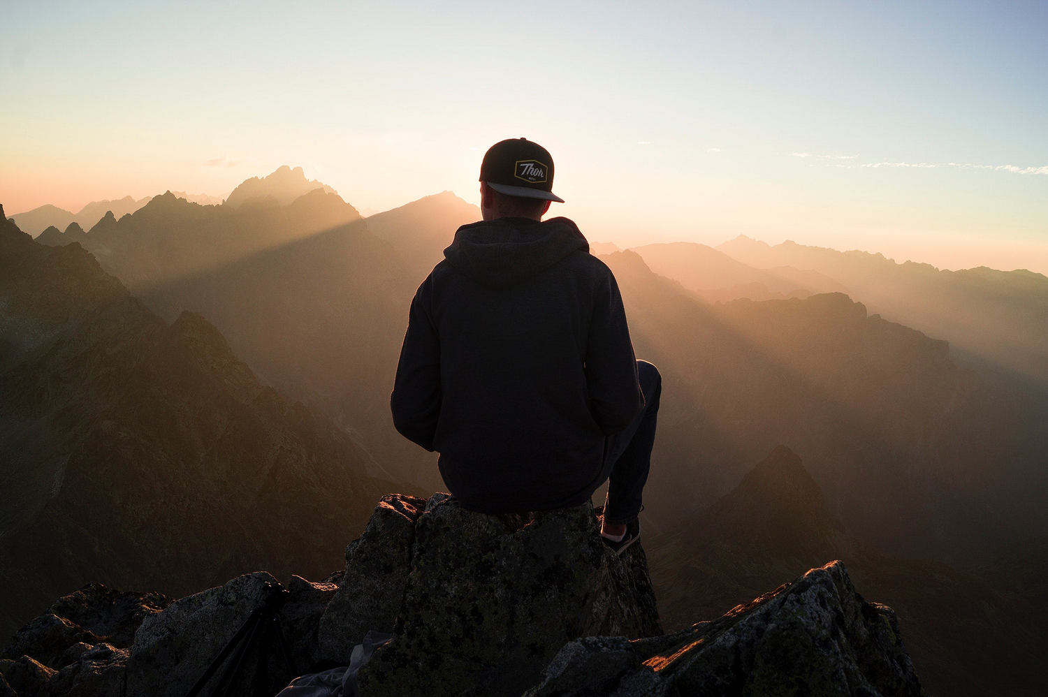 A young man sitting on top of a mountain rock, overlooking a mountain range